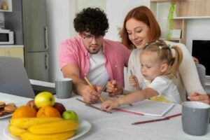 A family gathers around a table, engaged in a creative activity with colorful pencils, mugs, and a bowl of fruit nearby.