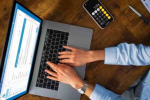 A person's hands typing on a laptop keyboard, with a calculator app open on their phone, set against a wooden desk.