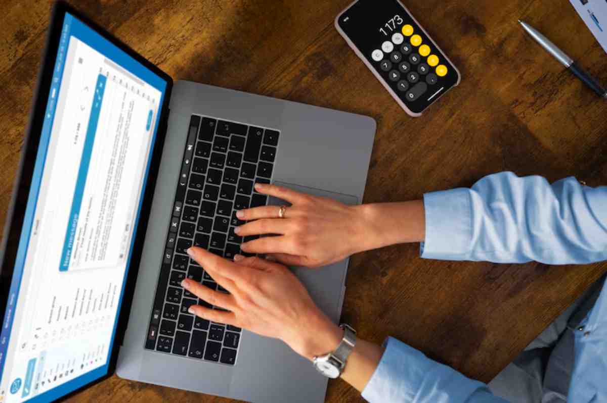 A person's hands typing on a laptop keyboard, with a calculator app open on their phone, set against a wooden desk.