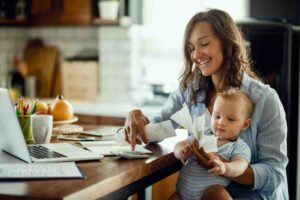 A woman works at a wooden table with a laptop, calculator, and receipts, holding a baby in her arms among kitchen items.