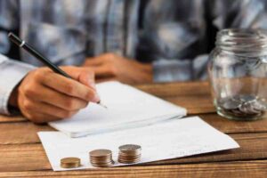 A person writes notes with a pen on paper, accompanied by stacks of coins and a jar filled with loose change on a wooden table.