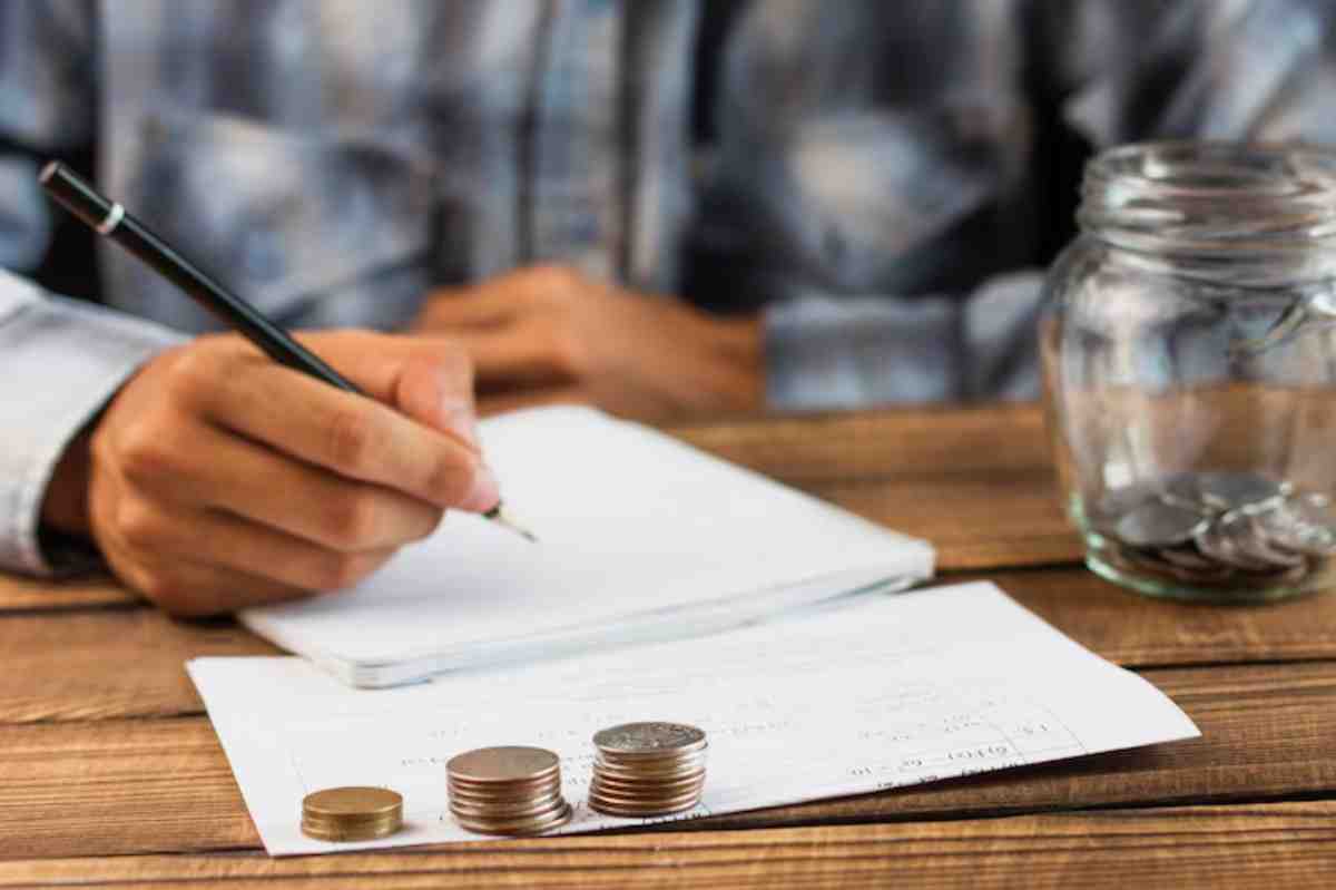 A person writes notes with a pen on paper, accompanied by stacks of coins and a jar filled with loose change on a wooden table.