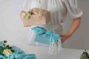 A person in a white dress holds two beautifully wrapped gifts: one in natural fabric with flowers and another in blue striped cloth.