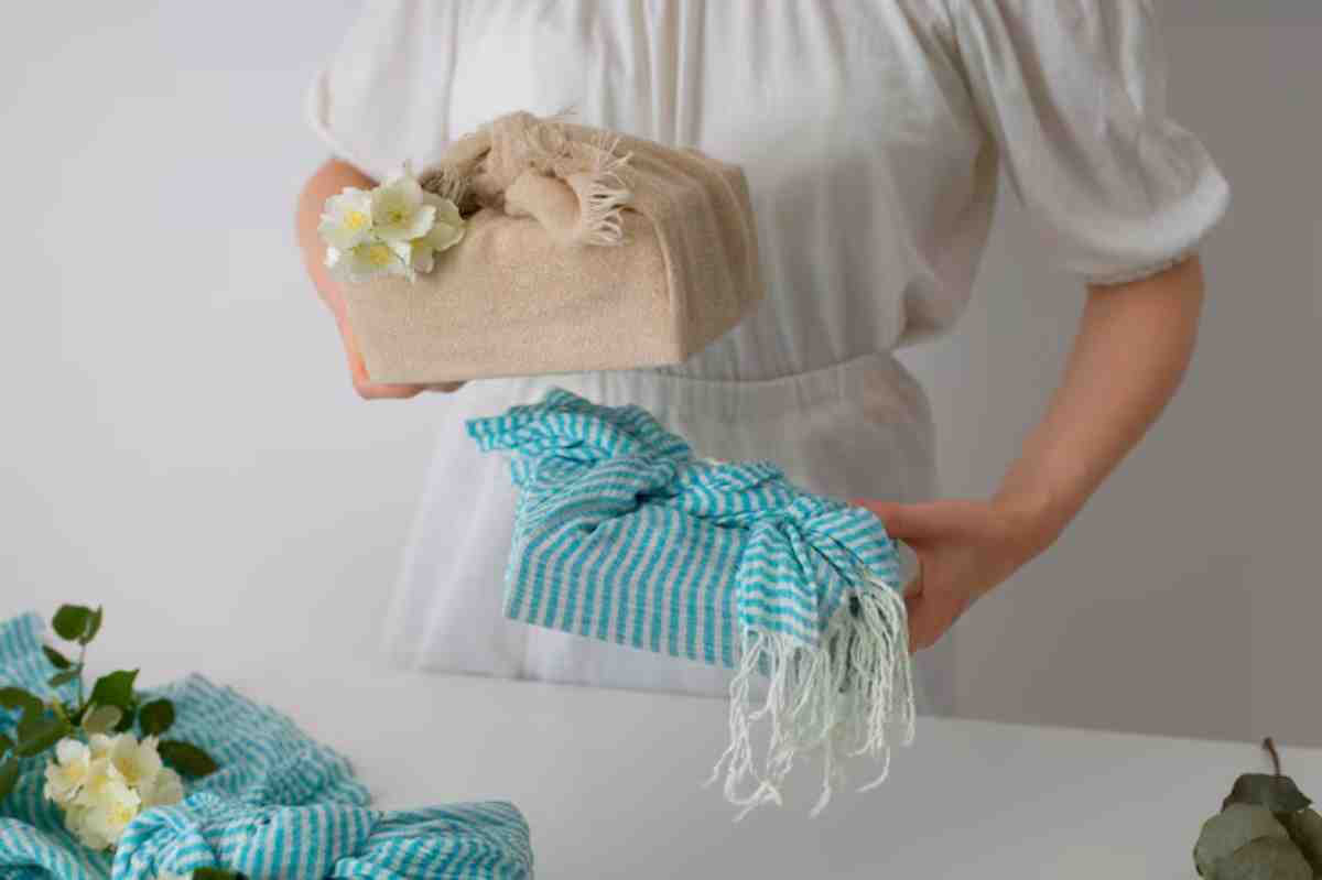 A person in a white dress holds two beautifully wrapped gifts: one in natural fabric with flowers and another in blue striped cloth.
