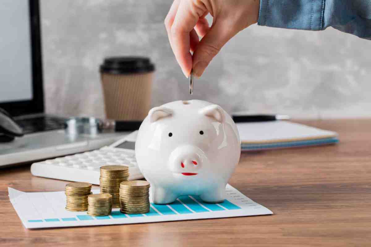 A hand drops a coin into a white piggy bank, surrounded by stacks of coins on a financial document and a laptop.
