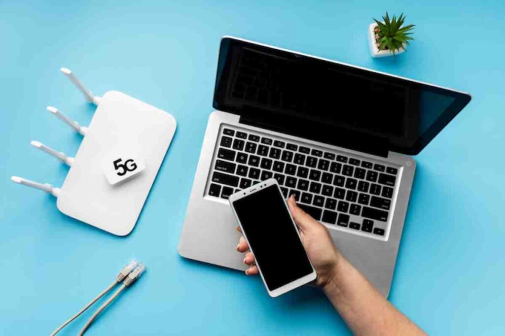 A hand holds a smartphone next to a laptop and a 5G router, set against a blue background with a small plant and Ethernet cables.
