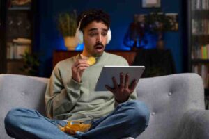 A person sits comfortably on a gray couch, wearing headphones, enjoying snacks from a bowl while using a tablet. Cozy ambiance with blue lighting.