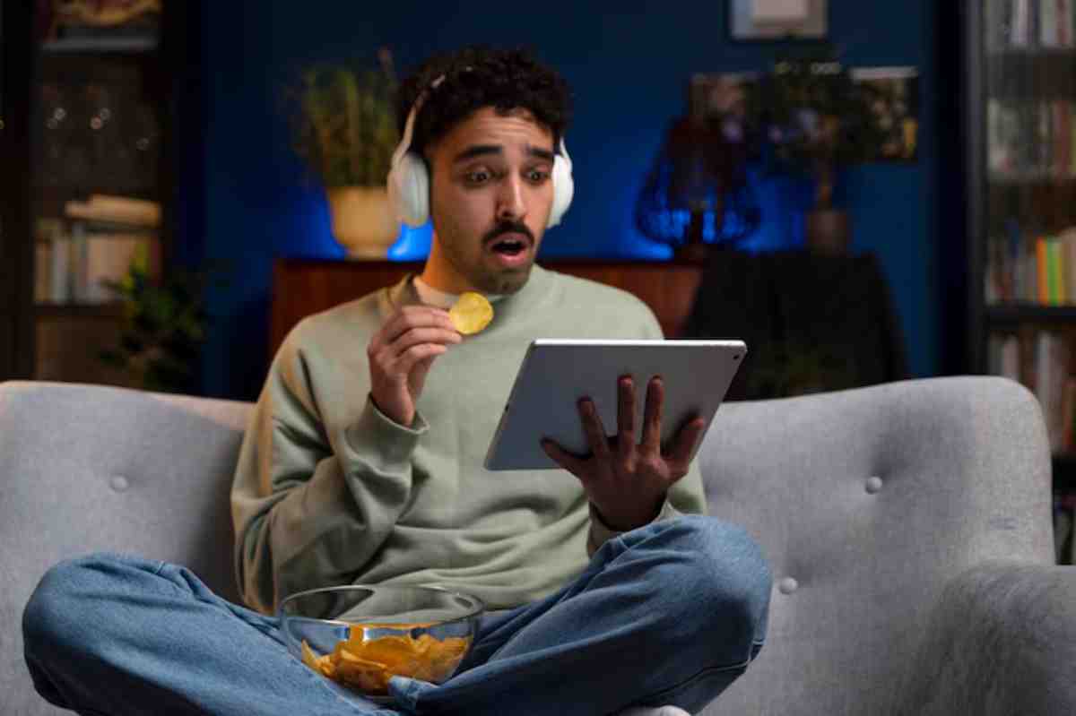 A person sits comfortably on a gray couch, wearing headphones, enjoying snacks from a bowl while using a tablet. Cozy ambiance with blue lighting.
