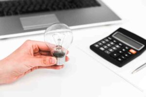 A person holds a light bulb above a laptop and a calculator on a white desk, symbolizing ideas and calculations.