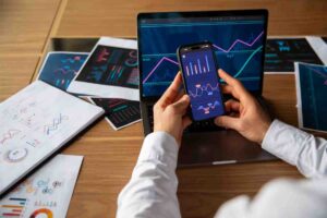 A person holds a smartphone displaying graphs, while a laptop shows similar data. Printed reports with visual data are scattered on a table.