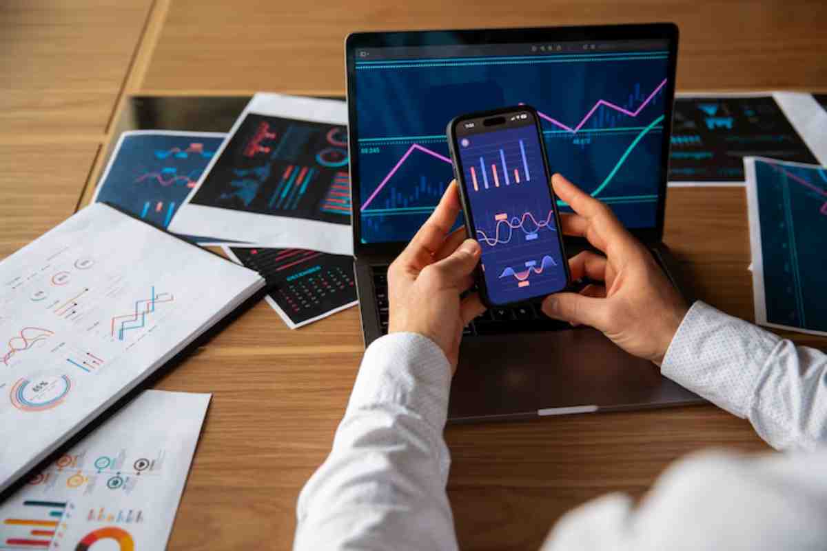 A person holds a smartphone displaying graphs, while a laptop shows similar data. Printed reports with visual data are scattered on a table.