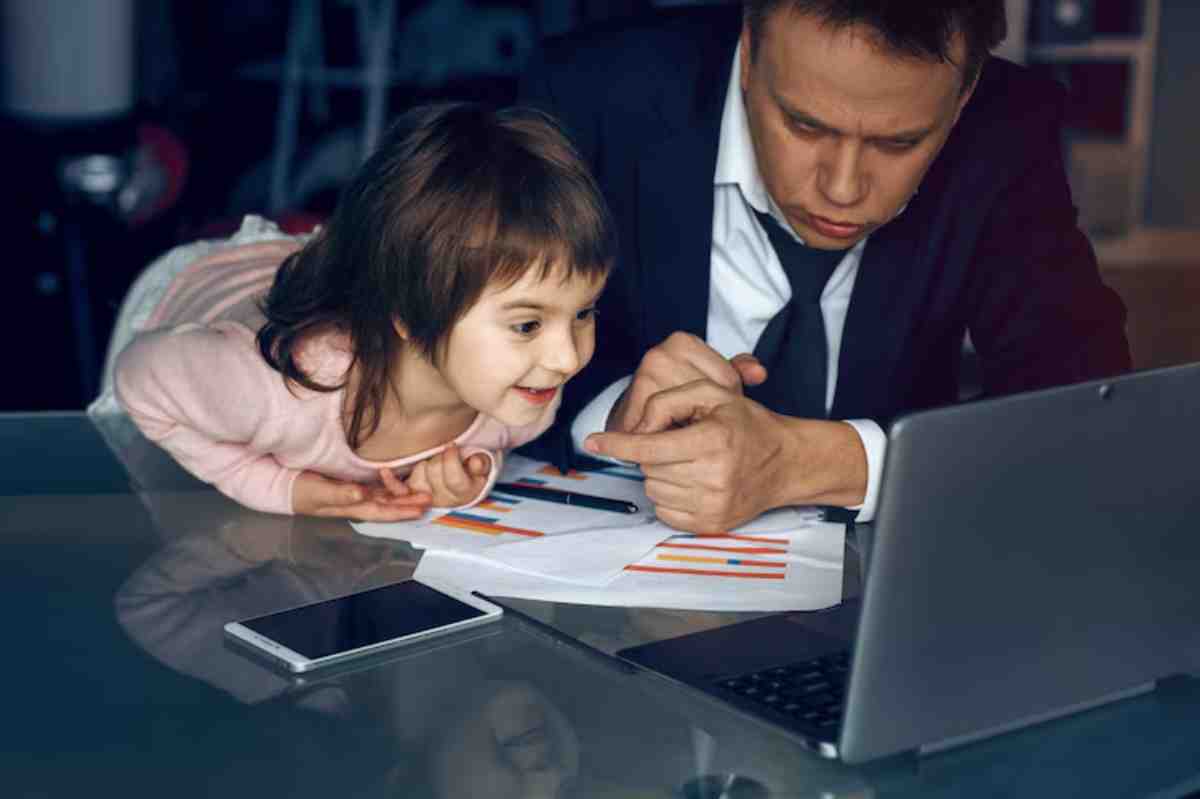 A man in a suit and a girl lean over a table with charts, discussing projects amidst a laptop and smartphone.
