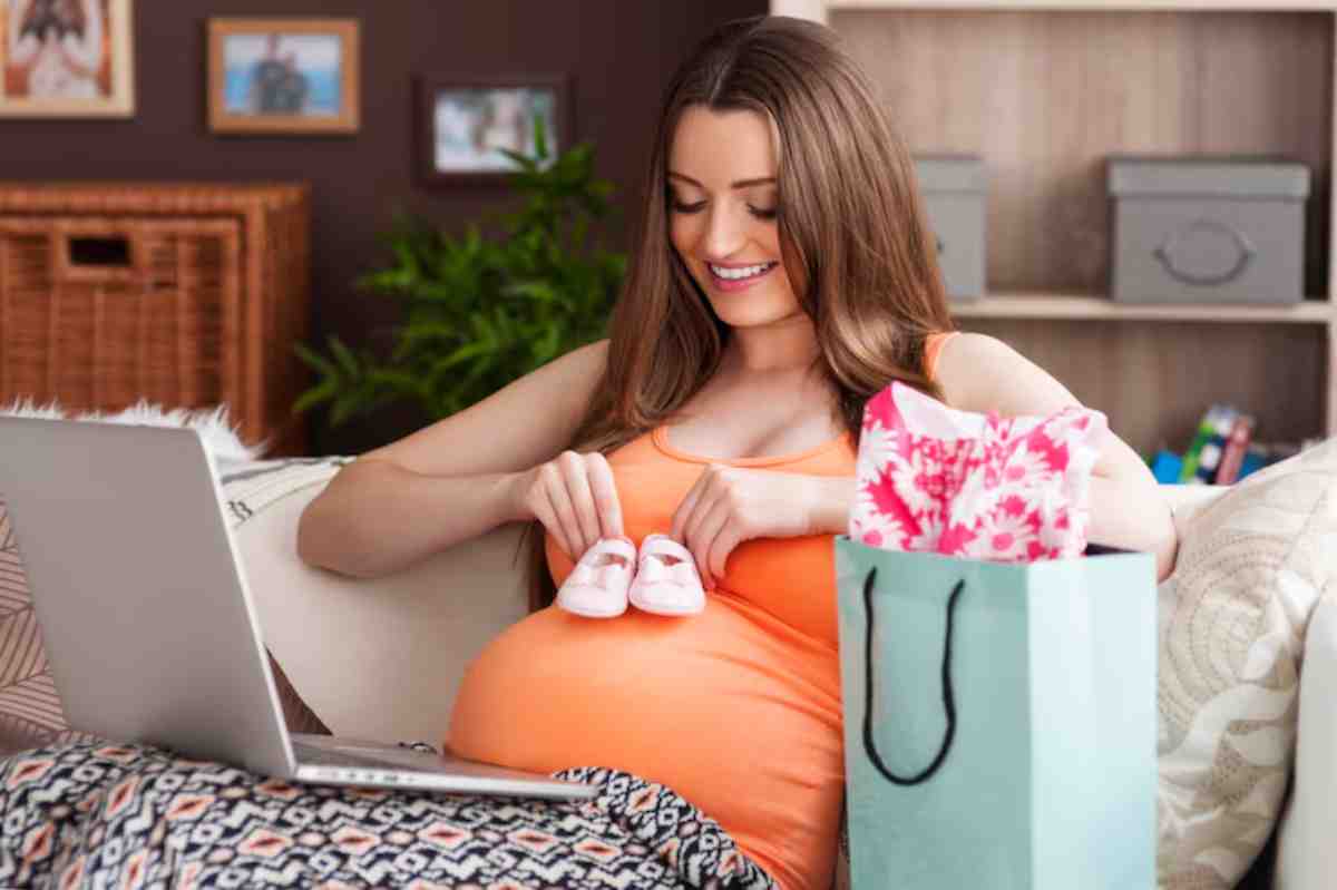 A pregnant woman in an orange top sits with a laptop, holding pink baby shoes, surrounded by gifts and cozy decor.