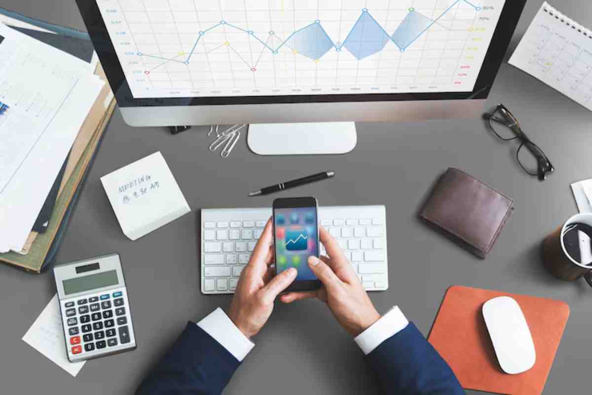 A person in a suit holds a smartphone displaying a graph, using a computer with a financial chart, calculator, and office supplies nearby.