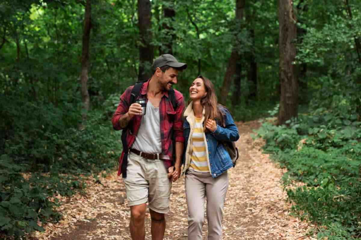A couple walks hand-in-hand along a leafy forest path, with trees and greenery surrounding them, creating a serene outdoor atmosphere.