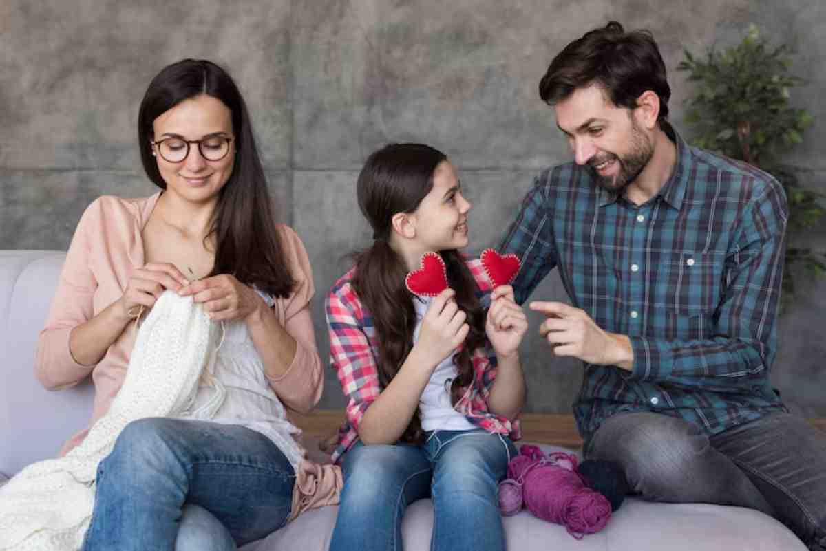 A woman knits while a girl and a man share cheerful moments, with the girl holding red fabric heart shapes. Colorful yarn is nearby.
