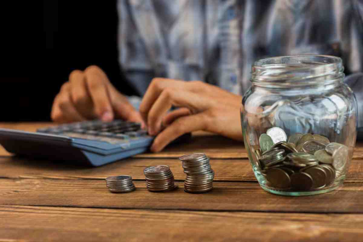 A person calculates finances using a calculator, with stacks of coins and a jar filled with coins on a wooden table.