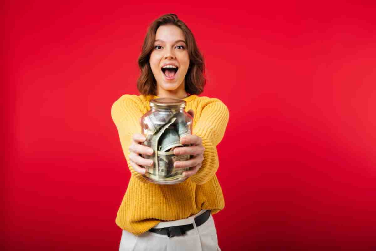 A person in a yellow sweater holds a glass jar filled with cash, set against a vibrant red background.