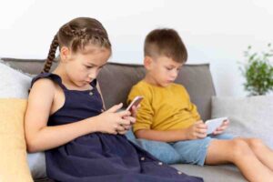 Two children sit on a couch, each focused on their smartphones, surrounded by soft pillows and a potted plant in the background.
