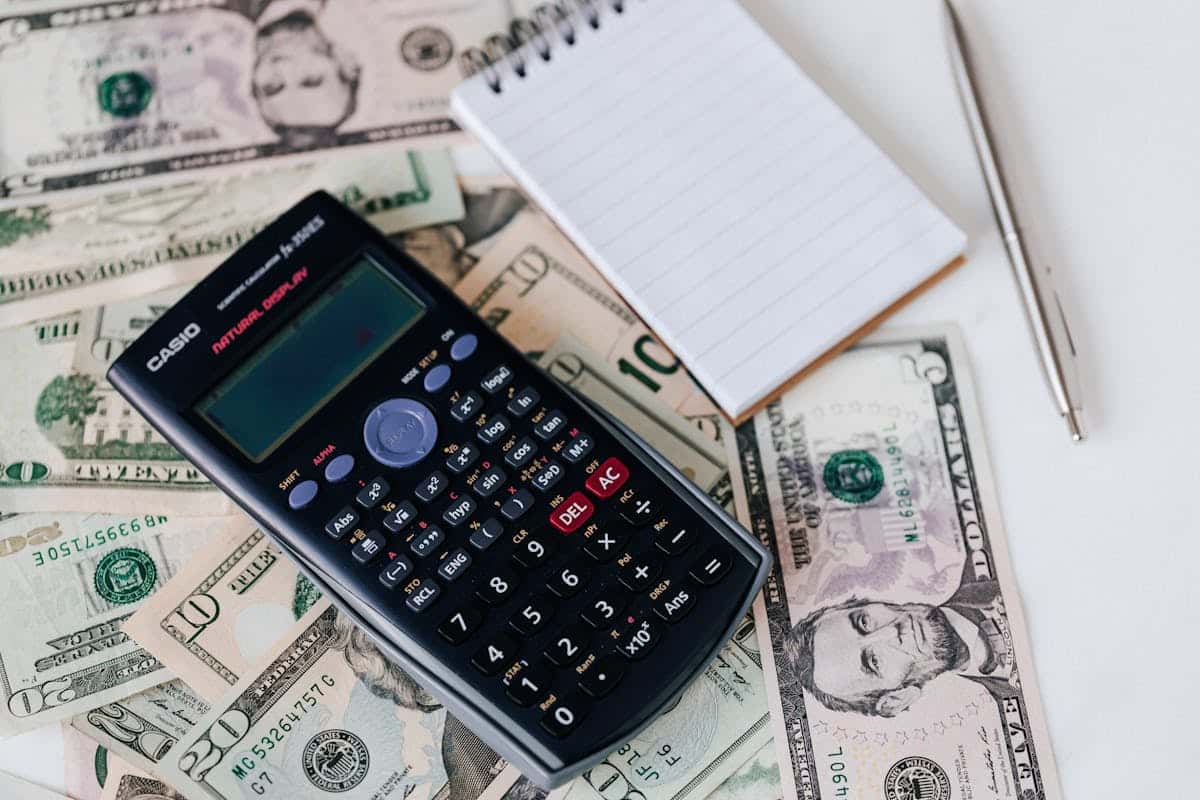 A calculator on a pile of US dollar bills with a blank notepad and pen nearby, symbolizing financial planning or budgeting.