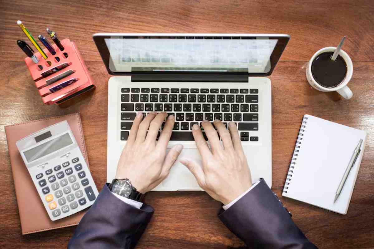 A person in a suit typing on a laptop, surrounded by a calculator, notepad, pen, coffee cup, and a pencil holder on a wooden desk.