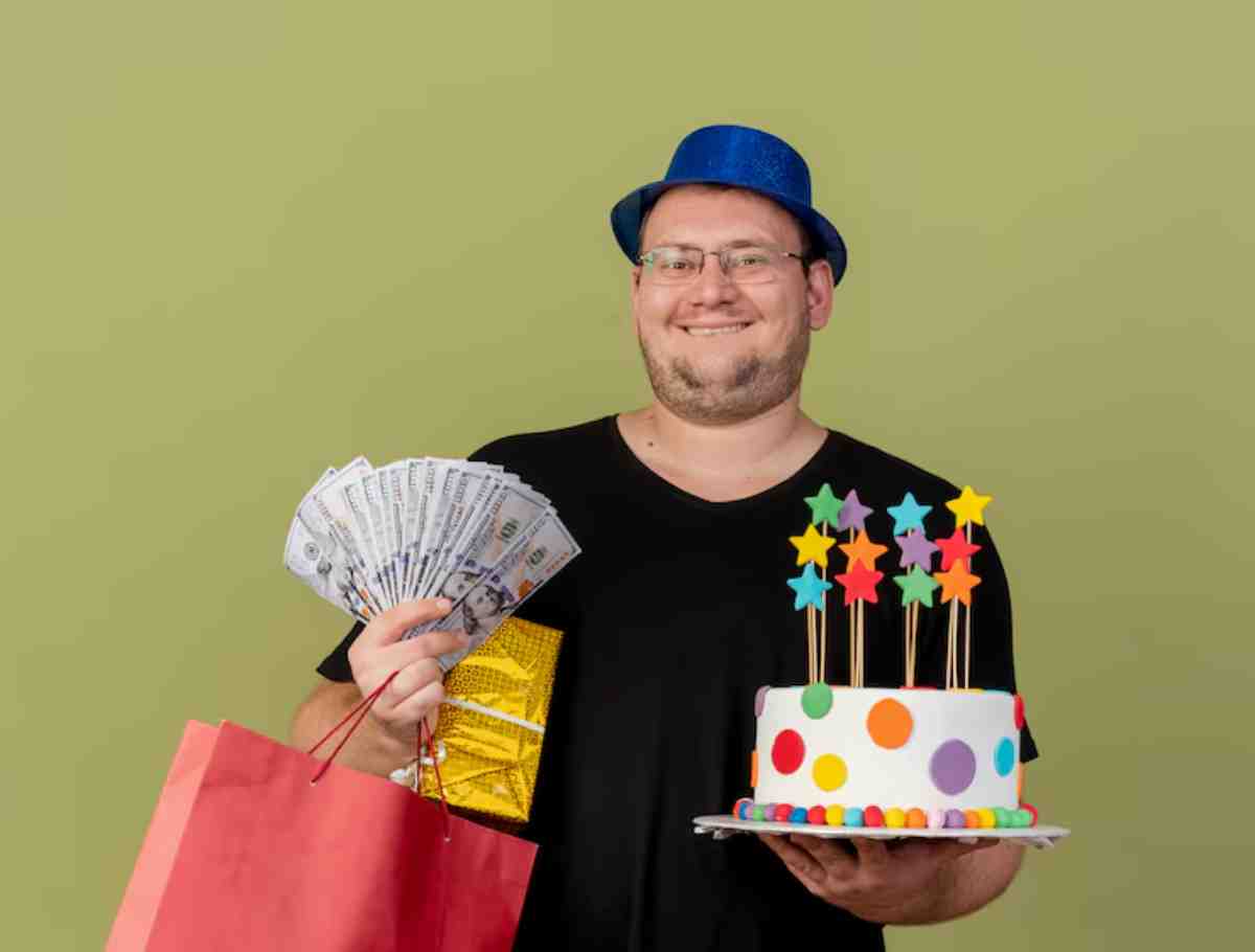 A person wearing a blue party hat holds a colorful cake with star decorations, money, and a gift bag against a green backdrop.