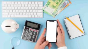 A person's hands hold a smartphone above a calculator, piggy bank, notepad, pencil, and euro banknotes on a blue background.