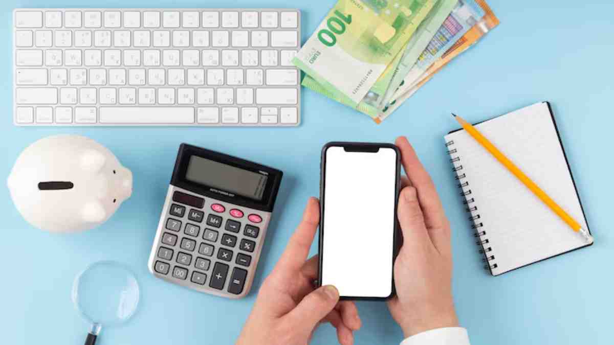 A person's hands hold a smartphone above a calculator, piggy bank, notepad, pencil, and euro banknotes on a blue background.
