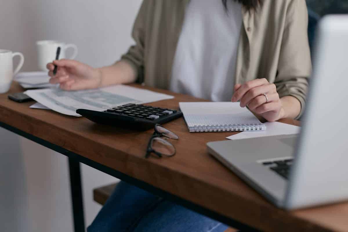 A person calculates finances at a wooden desk, surrounded by notebooks, papers, glasses, and two coffee mugs. A laptop is nearby.
