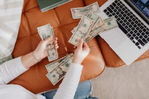 A person counts various U.S. banknotes while seated on a leather couch, with a laptop beside them.