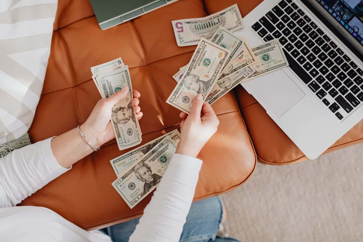 A person counts various U.S. banknotes while seated on a leather couch, with a laptop beside them.