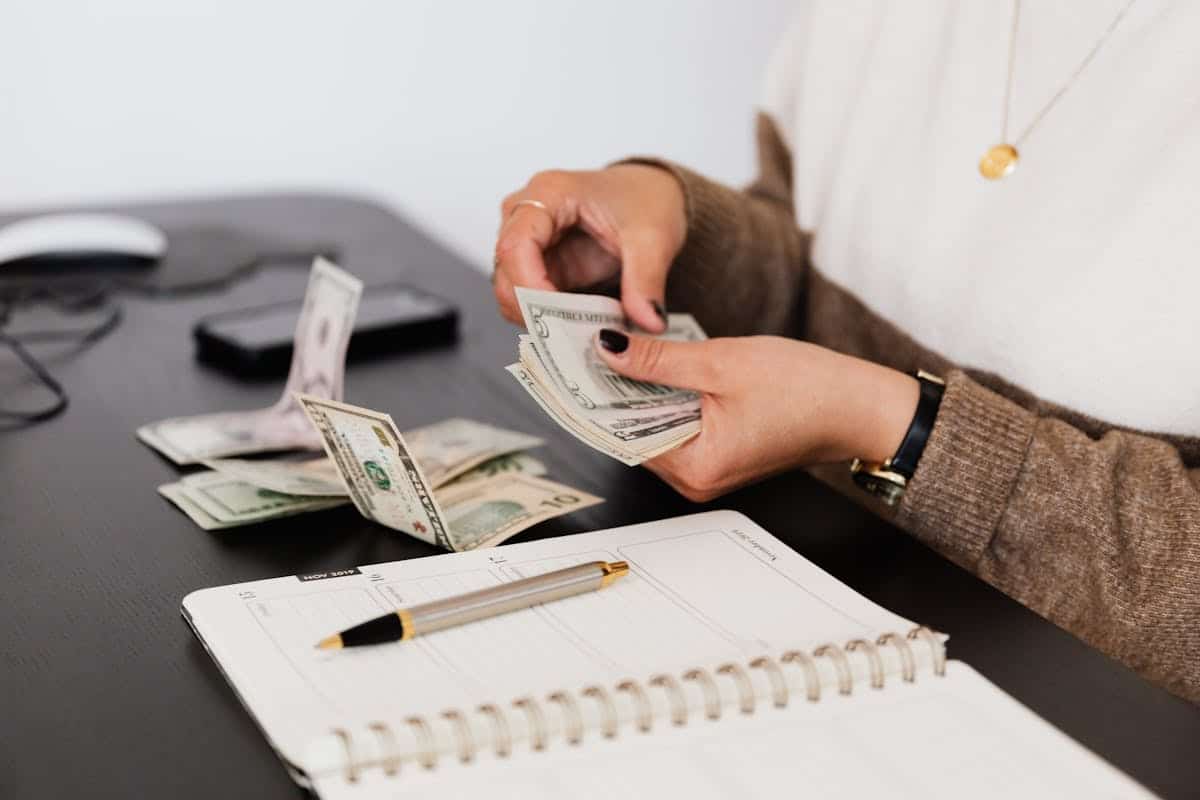 A person counts cash while seated at a desk with a planner and pen, a smartphone partially visible in the background.
