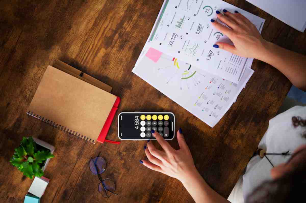 Top-down view of a person using a calculator on a smartphone, surrounded by graphs, a notebook, glasses, and a small plant.