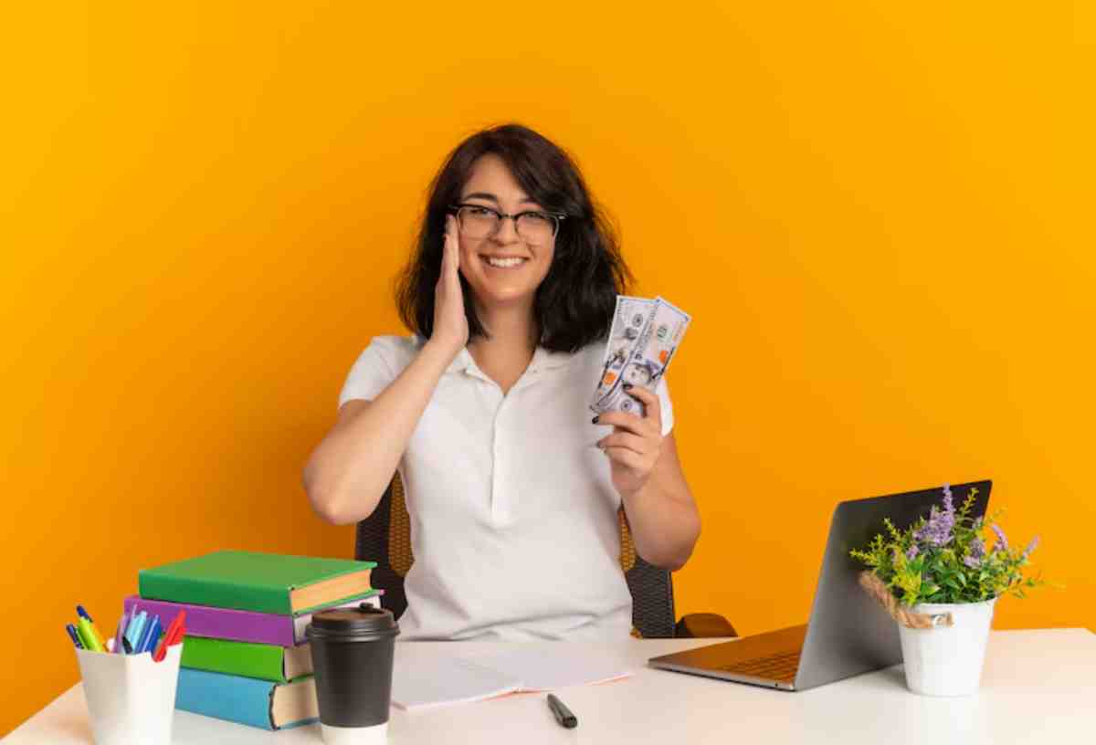 Person holding cash in one hand while seated at a desk with books, a laptop, a coffee cup, and a potted plant against a bright orange background.