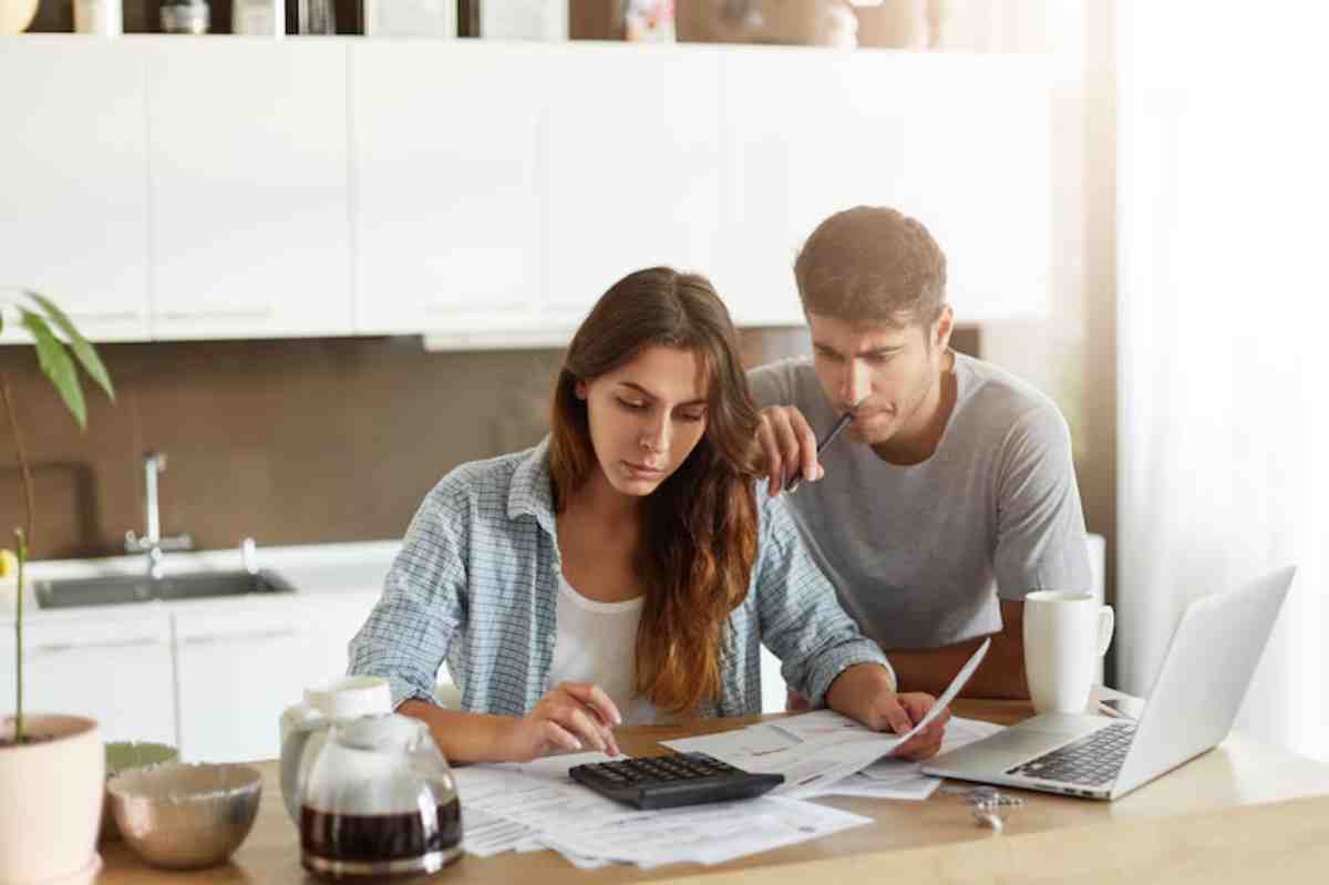 A couple reviews financial documents at a kitchen table, surrounded by a laptop, coffee, and papers, focused on budgeting.