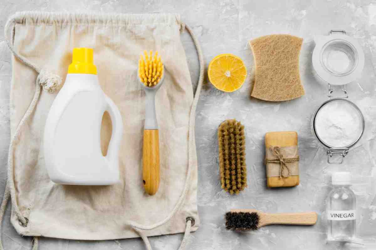 A collection of eco-friendly cleaning supplies: a bottle of cleaner, brushes, sponges, lemon, vinegar, and baking soda on a light surface.