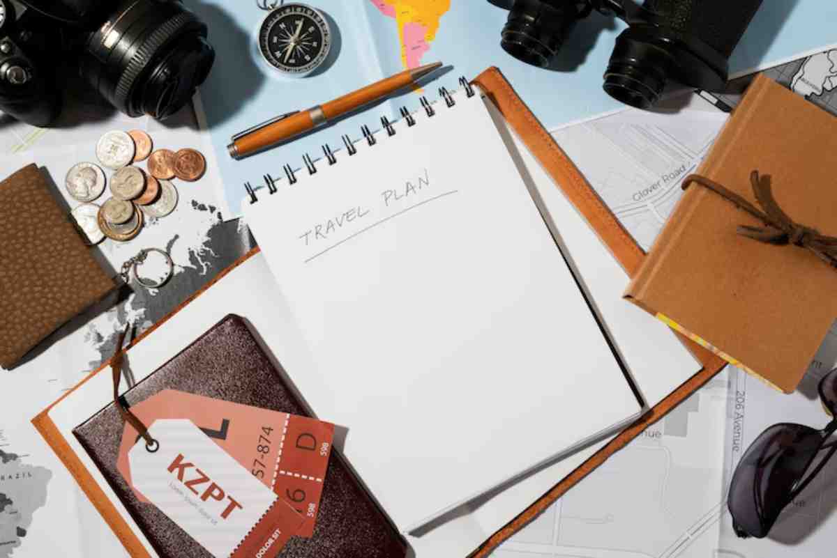A travel planning setup featuring a blank notebook, camera, compass, coins, maps, and travel tags, signifying adventure ahead.