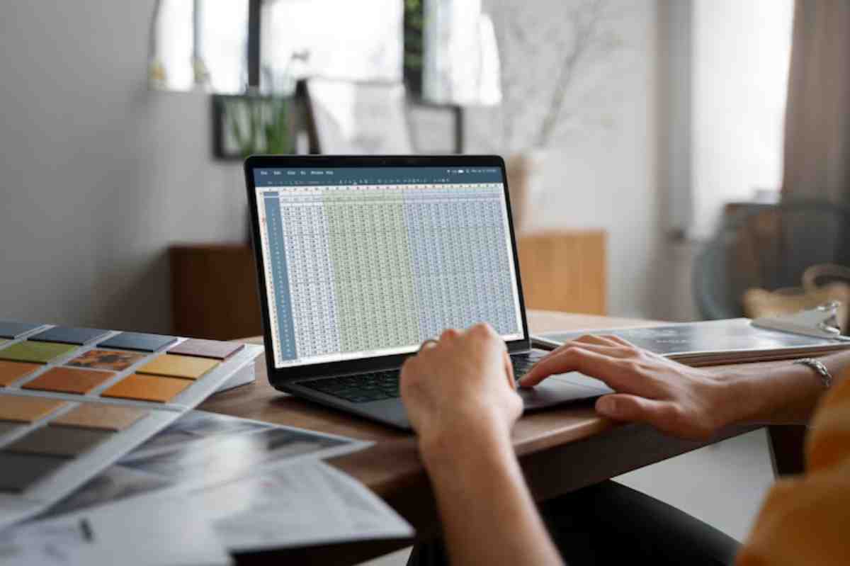 A person typing on a laptop displaying a spreadsheet, with color swatches and documents scattered on a wooden table.