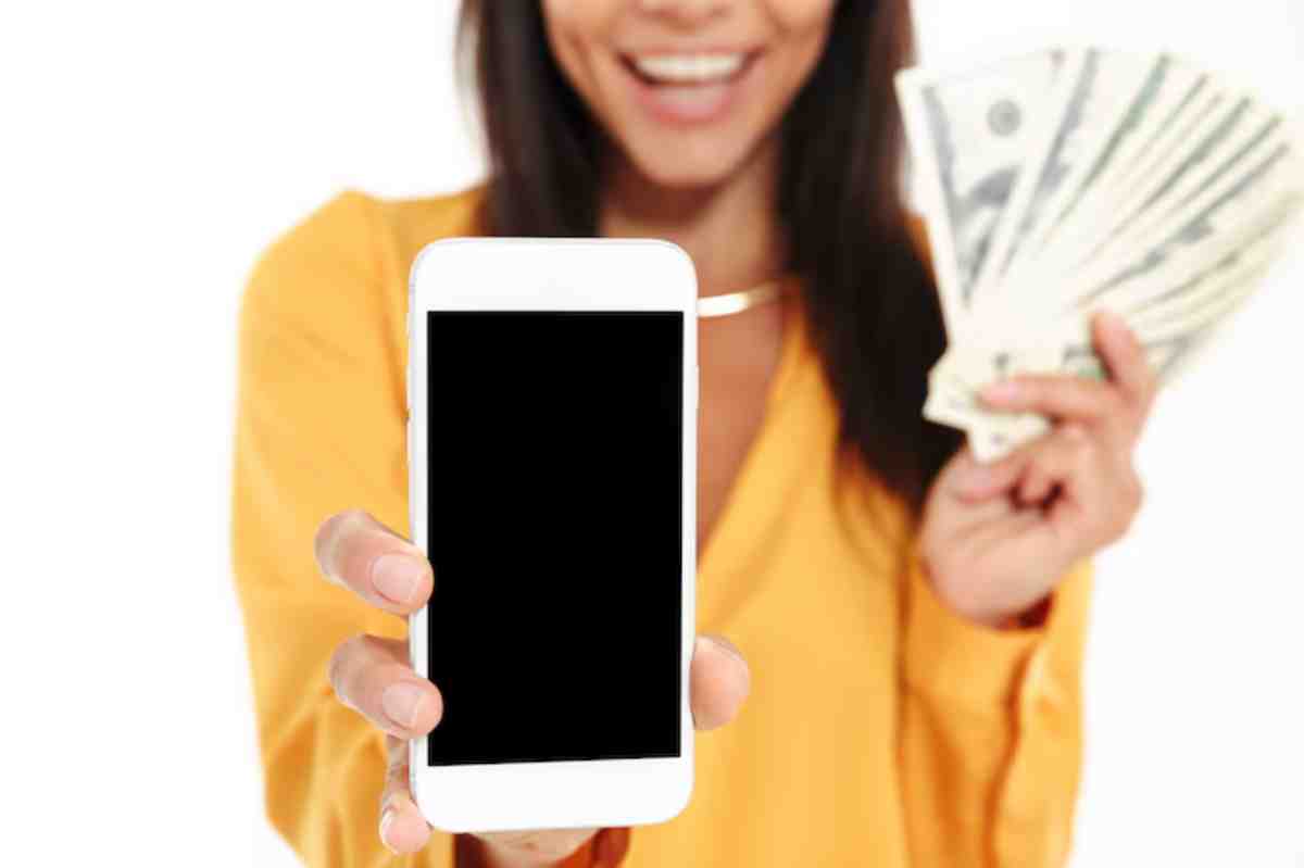 Smiling woman in a bright yellow shirt holds a smartphone and a fan of cash, symbolizing success or financial gain.