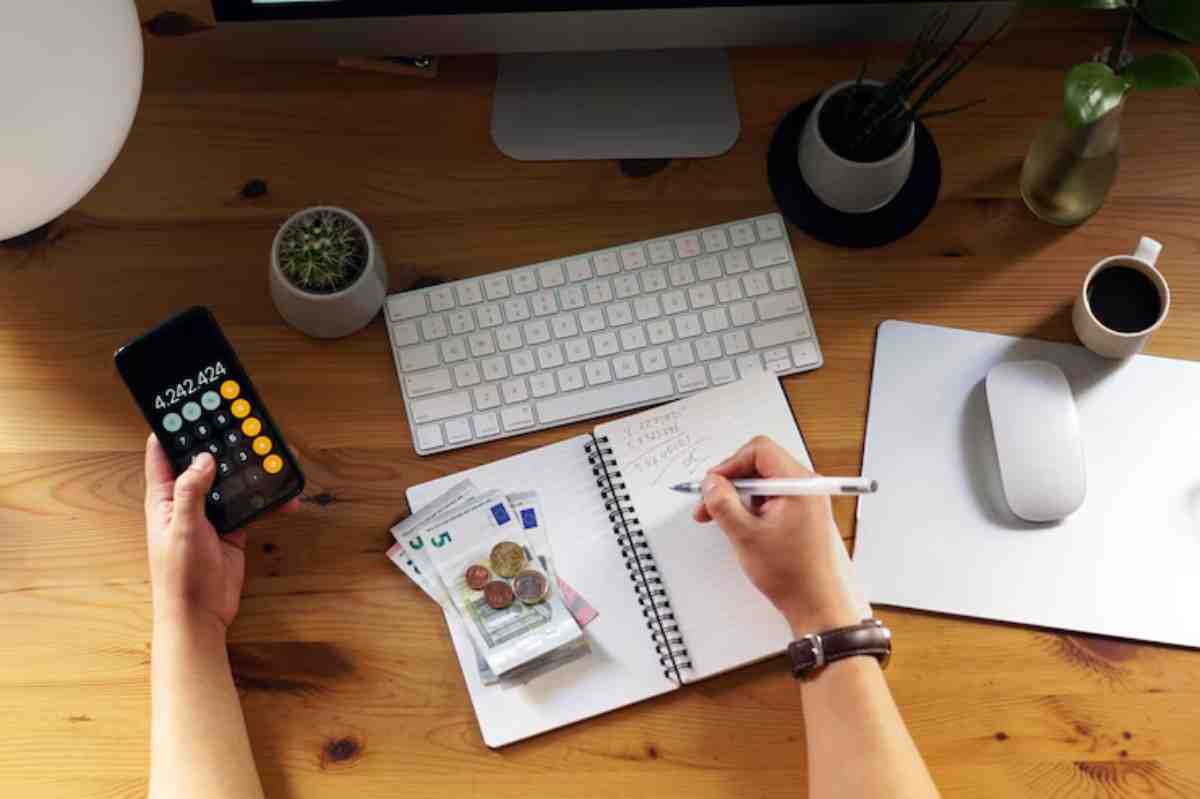 A person calculates finances on a smartphone, writes notes in a notebook, and has cash on a wooden desk with a computer and plants.