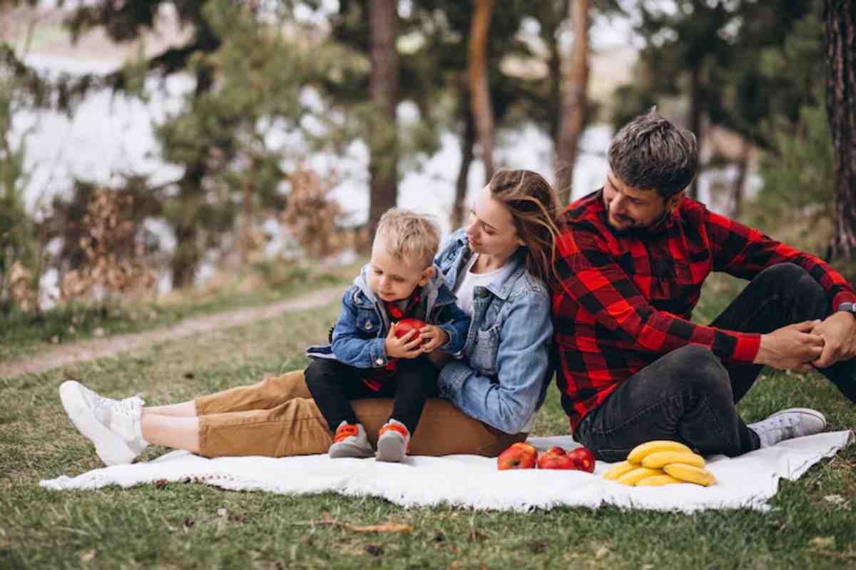 A family enjoys a picnic on a blanket, surrounded by trees, with bananas and apples arranged on the ground.