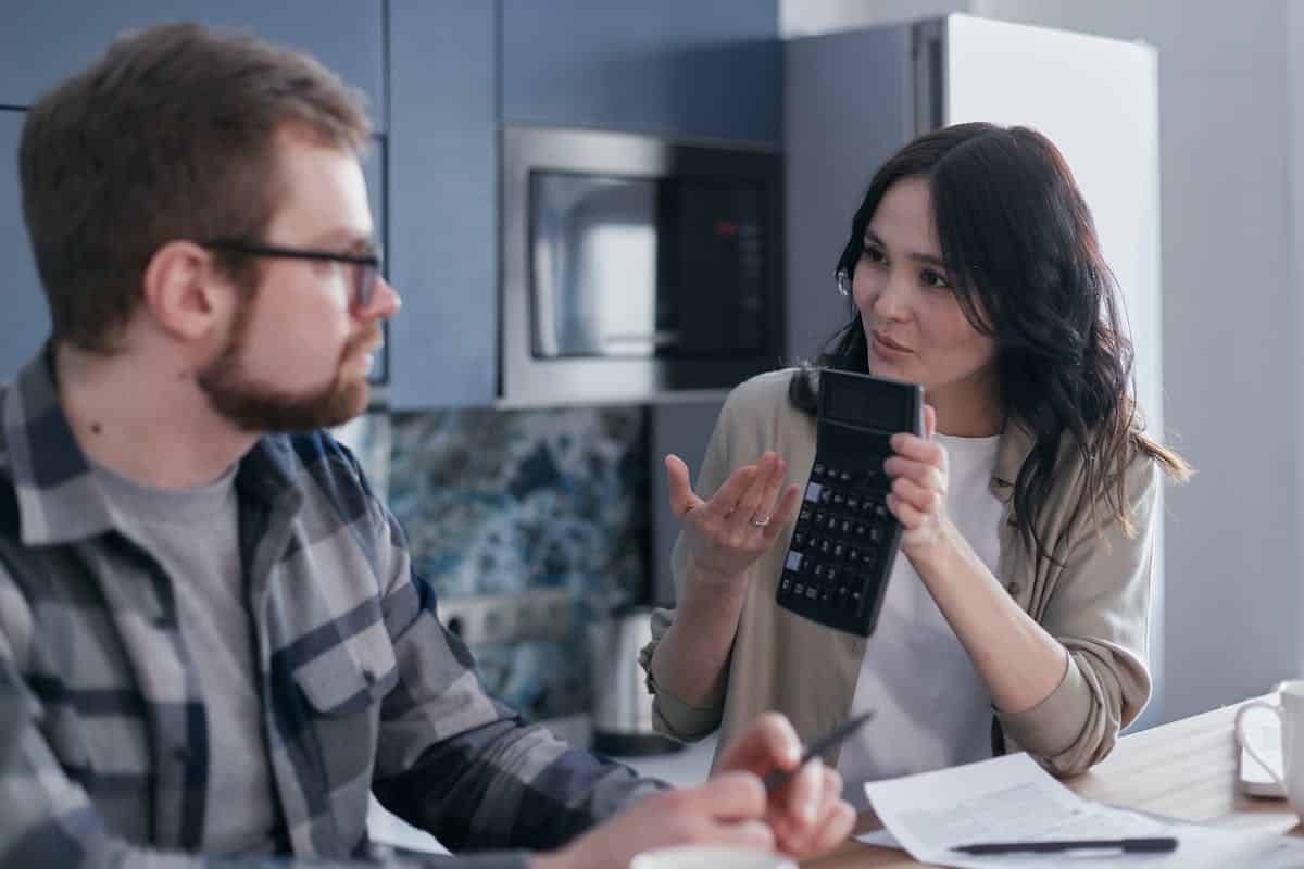 A woman in a beige cardigan holds a calculator and gestures while discussing with a man at a kitchen table covered with papers.