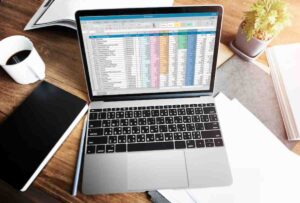 A laptop displaying a colorful Excel spreadsheet sits on a wooden desk alongside a coffee cup and a notebook.