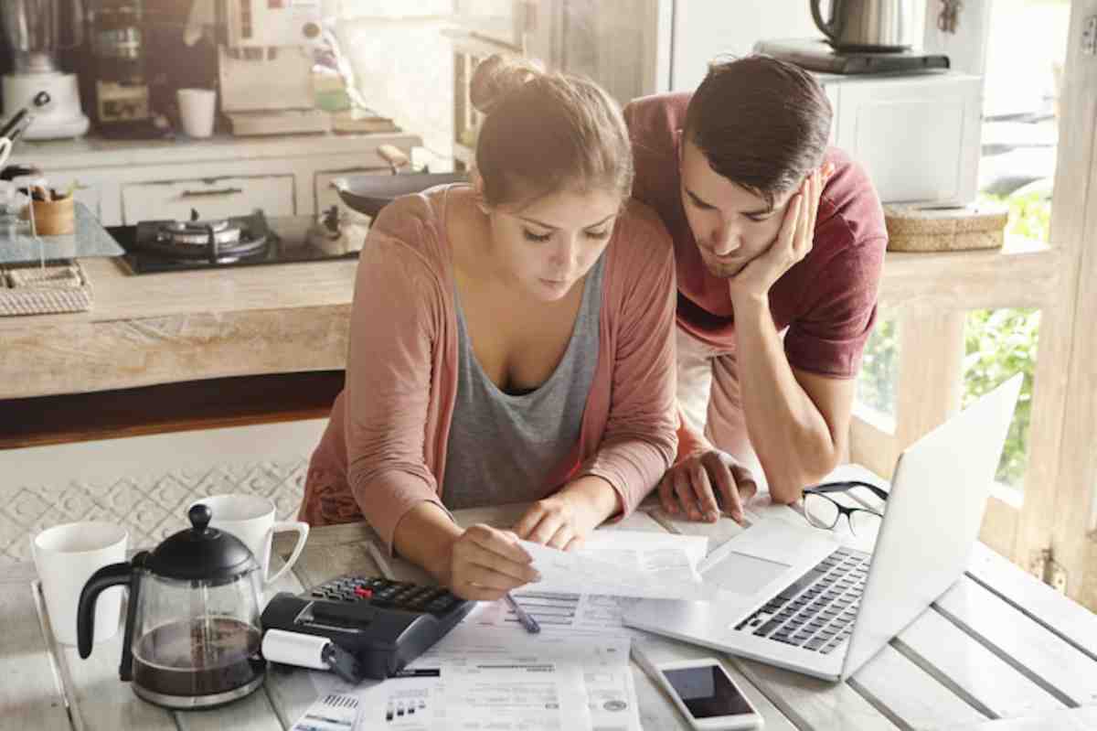 A couple studies financial documents at a wooden table, surrounded by coffee, a laptop, and a calculator in a cozy kitchen setting.