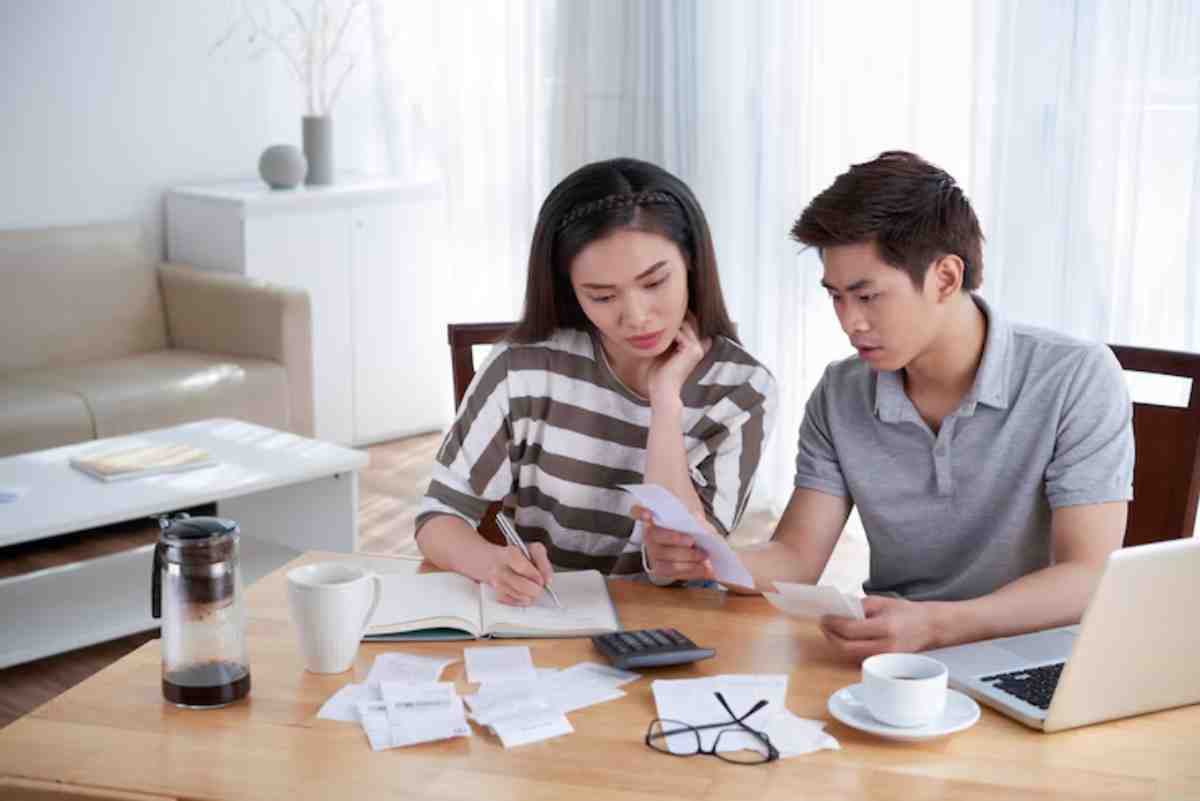 A couple sits at a wooden table, reviewing documents, surrounded by coffee, a calculator, and scattered receipts in a bright room.