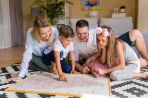 A family gathers around a map on a rug, engaged in discussion, with a warm and cozy living room setting in the background.