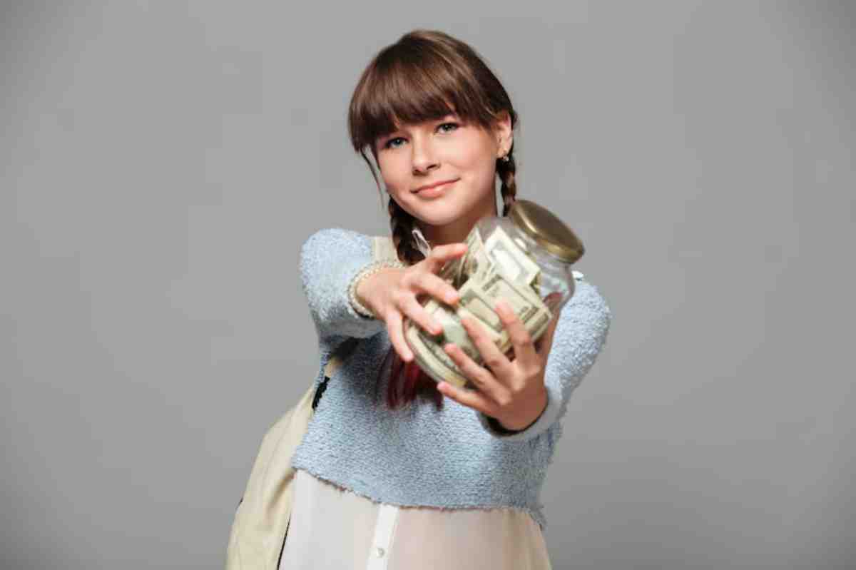 A person in a light blue sweater holds out a glass jar filled with money, displaying excitement against a gray background.