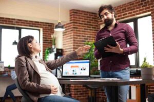 A pregnant woman sits at a desk gesturing, while a man stands beside her holding a clipboard, both engaged in discussion in a modern office.