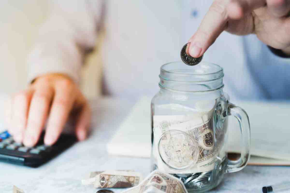 A person drops a coin into a jar filled with cash while using a calculator, symbolizing saving and budgeting.