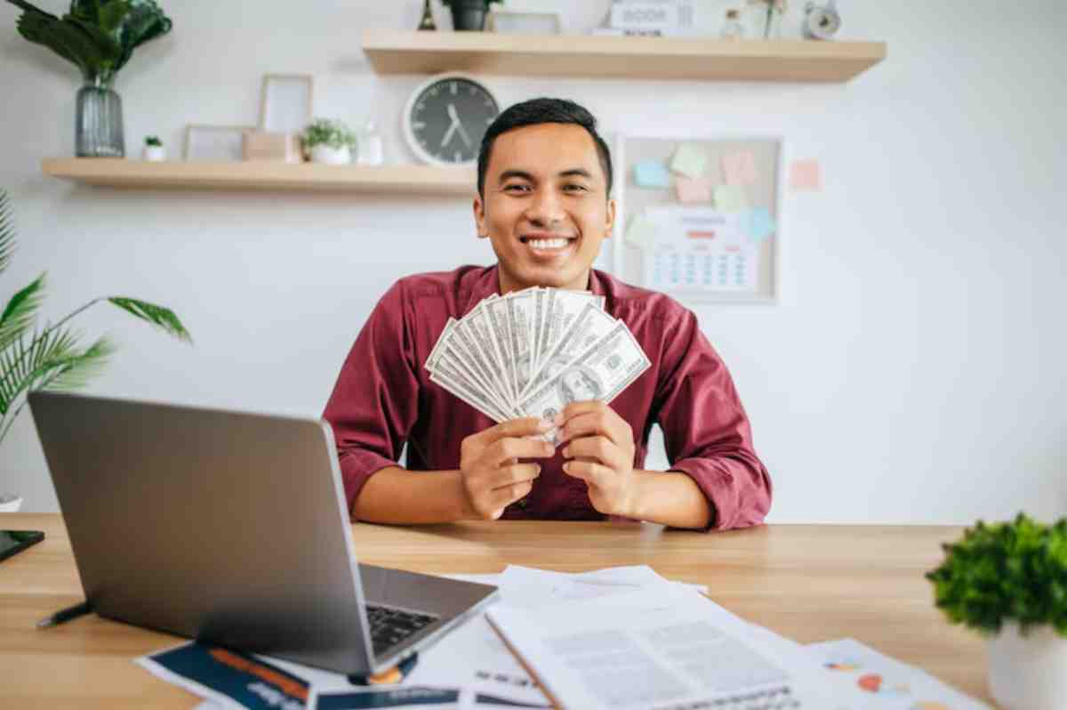 A person in a maroon shirt holds a fan of hundred-dollar bills in front of a laptop and paperwork on a wooden desk.